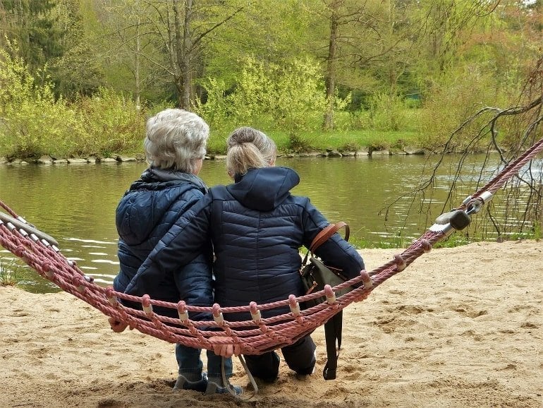 This shows an older lady and a middle aged lady sitting on a hammock by a lake