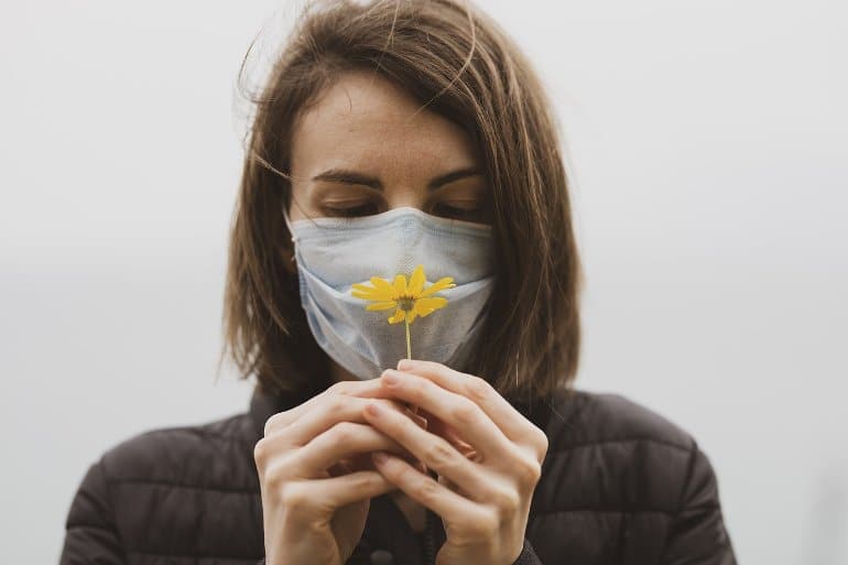 This shows a woman in a facemask holding a flower to her nose