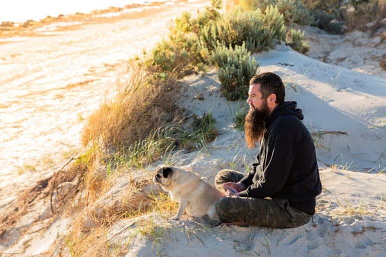 This shows a man sitting on a beach with his dog