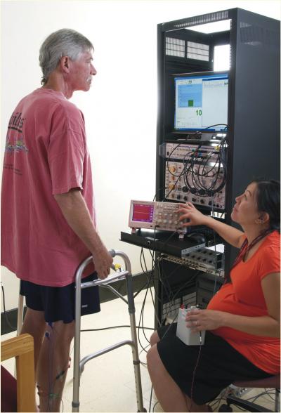 A man is standing with the help of a walker in front of a tower of electronics as a woman sits nearby.